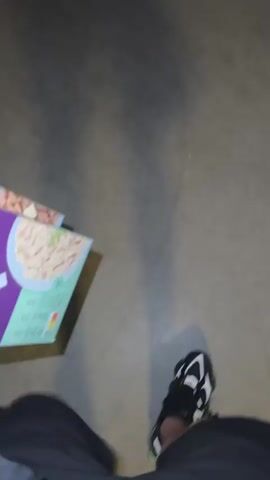 A first-person perspective video shows someone walking while holding two boxes of "Fine Barley." The boxes are purple with images of the barley dish on them. The person is wearing black shorts and black and white Adidas shoes with green accents. The floor is a dark, smooth surface, possibly concrete. The lighting is dim.