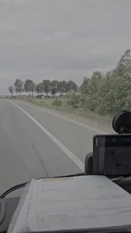 The video is taken from the perspective of someone inside a vehicle, likely a truck or a car, driving on a highway. The view is directed forward, showing the road ahead and the scenery. The road is paved and has a white line marking the lane. To the right of the road, there is a grassy area and a line of trees. The sky is overcast and appears to be a cloudy day. In the foreground, there is a dashboard with some papers and a navigation device or phone mounted. The vehicle is in motion, indicated by the perspective and the blurred road markings. There are no people visible in the video. The overall scene depicts a journey on a highway under a cloudy sky.