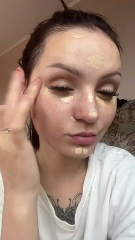 The video shows a close-up of a woman applying makeup. She has dark hair pulled back and is wearing a white shirt. Her face is dotted with what appears to be foundation or concealer, particularly under her eyes, on her cheeks, and across her forehead. She is using her fingers to blend the product, focusing on the area under her left eye. The lighting is soft, and the background is blurred, suggesting an indoor setting.
