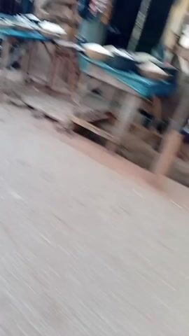 The video shows a street in an outdoor setting, likely a village or a rural area. The camera moves along the road, capturing the view of the side of the street. Small buildings and structures, possibly shops or houses, line the road, constructed from materials like wood and corrugated metal. A person on a motorcycle passes by the camera. The overall atmosphere suggests a simple, everyday scene in a less urbanized area.