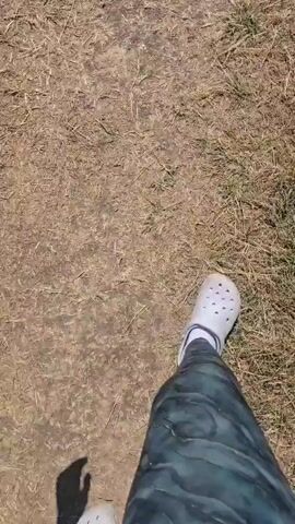 The video shows a first-person perspective of someone walking outdoors. The person is wearing white Crocs and dark, patterned leggings. The ground is a mix of dry dirt and sparse, brown grass. The camera follows the person's feet as they move forward. The lighting suggests it is daytime with sunlight.