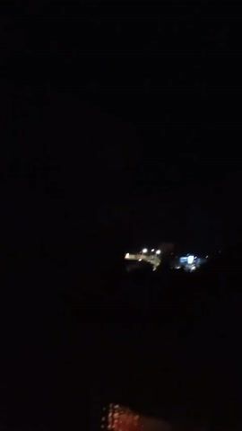A night view of distant buildings with illuminated windows and streetlights. The scene is dark with the lights of the buildings being the primary focus. The camera appears to be moving slightly, causing a subtle blur.