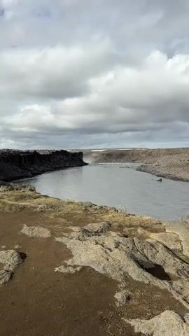 The video presents a static wide shot of a river flowing through a rugged, rocky canyon under an overcast sky, with a slight pan to the right and back. No people or specific objects are visible beyond the natural landscape.

The scene features a wide, grey river occupying the center of the frame, bordered by dark, imposing rocky canyon walls on both sides. These walls appear to be composed of layered or columnar basalt, characteristic of volcanic landscapes. The immediate foreground consists of dark, barren earth and scattered rocks with sparse, dry vegetation.

The river in the foreground is calm, but in the distance, it narrows and appears to accelerate towards a powerful waterfall, which is partially obscured by the canyon's bend. The audible rush of water reinforces the presence and power of the waterfall. The sky is completely covered with heavy grey clouds, creating a diffused light that emphasizes the rugged textures of the landscape. The time of day is clearly daytime, but the lack of direct sunlight gives the scene a slightly muted and dramatic appearance.

The overall impression is one of a vast, untamed natural environment, devoid of human activity. The rugged terrain, dark rocks, and powerful river suggest a remote location, consistent with the provided context of Iceland. No visible text is present in the media.