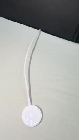 A white Apple Watch charging puck with a white cable is on a light-colored textured surface. A hand enters the frame from the right and places a silver Apple Watch with a gray band onto the charging puck. The watch is then centered on the puck.
