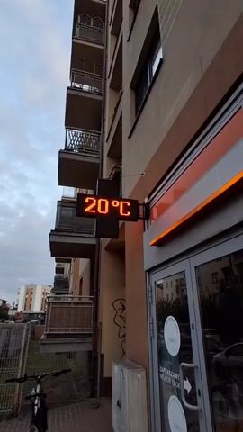 The video captures an exterior shot of a building, likely a pharmacy, as indicated by the illuminated sign that reads "APTEKA". The sign is an LED display showing the text "20°C" at the beginning of the clip, which then transforms into the word "APTEKA". The building appears to be a residential or commercial structure with balconies visible on its facade. An orange illuminated awning is above the entrance. A bicycle is parked near the entrance. The time appears to be late afternoon or early evening due to the lighting conditions.