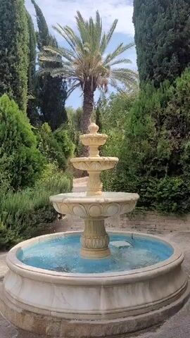 The video shows a multi-tiered fountain in a garden. The fountain is made of light-colored stone with decorative carvings. Water cascades from the top tier to the bottom basin, which is filled with blue-tinted water. Surrounding the fountain are lush green bushes, cypress trees, and a palm tree. The ground is paved with stone. The sky is visible and appears to be partly cloudy. The camera pans from left to right, showing different angles of the fountain and the surrounding garden.
