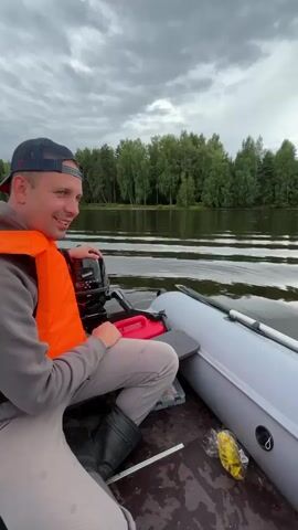 A man wearing an orange life vest and a baseball cap sits in an inflatable boat on a body of water. He is smiling and looking to his right. Behind him is an outboard motor and a red gas can. To his right, another person's hand is holding a slice of bread, offering it to him. The water is calm with ripples. In the background, a treeline is visible under a cloudy sky. The boat appears to be moving or about to move, as evidenced by the ripples in the water and the presence of the motor.