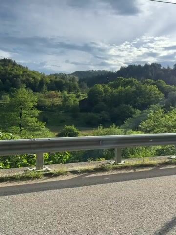 A car travels along a winding asphalt road through a mountainous, forested region. The sky is partly cloudy with the sun shining brightly. The vehicle passes a roadside guardrail on the right. Trees line both sides of the road. The interior of the car is briefly visible, showing the steering wheel and dashboard. The sun glares on the windshield, obscuring some detail.