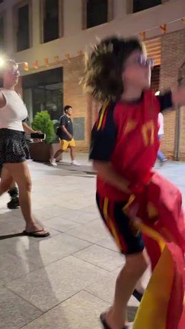 A young person, wearing a red and yellow Spanish national team jersey with the number 17 visible and dark shorts with red and yellow stripes, is in motion, holding a Spanish flag which is partially visible and unfurled. Their hair is long and appears to be moving due to their activity. They are also wearing glasses. The setting appears to be an outdoor urban environment, possibly a street or plaza at night, with brick buildings and string lights visible in the background. Other individuals are present in the background, one of whom is wearing a similar Spanish team jersey and holding a flag. The overall atmosphere suggests a celebration or event, possibly related to a sporting match. The ground is paved with light-colored stones. There is a slight motion blur, indicating movement within the frame.