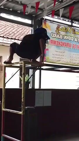 A person in a dark shirt and shorts is performing a difficult physical maneuver on a metal bar setup. They are crouched with their hands on one bar and their feet on another, their body angled in a way that suggests significant strength and balance. They are wearing a blue cap. The background shows a tiled roof, some plants, and a banner with text in what appears to be Indonesian. The overall environment seems to be an outdoor or semi-outdoor space, possibly associated with a school or community center, given the banner and the presence of play equipment. The lighting is natural daylight.