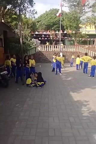 The video shows a group of children in matching yellow shirts and blue pants with yellow stripes on the sides, gathered in an outdoor paved area. Some children are standing, while others are sitting on the ground. In the background, there is a building with orange and yellow walls, and a staircase. There are also flags and triangular pennants strung up. A flagpole stands prominently in the foreground. The children appear to be engaged in some sort of activity or waiting to participate in an event. The lighting suggests it is daytime, and the weather appears to be clear.