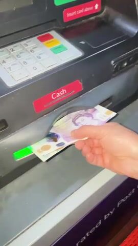 A close-up shot shows a person's hand withdrawing money from an ATM. The ATM is dark grey/black with a silver numeric keypad on the left, featuring 'CLEAR', 'CANCEL', and 'ENTER' buttons. Above the keypad, a red label with an upward arrow points to a slot, stating "Insert card above".

The hand is pulling a £20 banknote from a slot labeled "Cash", which has a glowing green light inside. The banknote is purple and yellow, clearly showing the portrait of Queen Elizabeth II and "Bank of England", "TWENTY POUNDS". Below the cash slot, partially visible text reads "generated by Post O", suggesting the ATM is located at a Post Office. The background is a grey wall or structure. The video captures the moment the money is being retrieved from the machine.