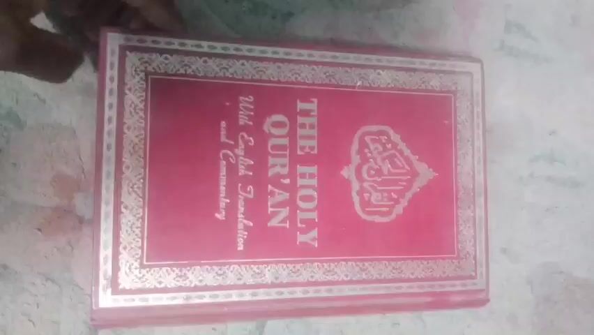 The video captures a close-up view of a book being opened and pages being flipped. A hand, appearing to be a person of color, is visible turning the pages of the book. The book has a pink cover on the right side and a white interior. The pages contain text in what appears to be Arabic script. The background is blurred and appears to be a textured surface, possibly a carpet or fabric. The lighting is dim, casting shadows and giving the scene a somewhat hazy quality. The action is focused on the book and the act of turning pages, suggesting an exploration or reading of the book's contents.