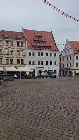 This video captures a panoramic view of a cobblestone town square in Pirna, Germany. The scene is characterized by charming, historic architecture, with buildings featuring red-tiled roofs and ornate facades. Colorful bunting, made of triangular flags in red, yellow, and orange, is strung across the square, suggesting a festive occasion or event.

Several buildings are visible, including a prominent one with a tall clock tower that has a green spire. This building appears to be a significant landmark. Another building has a coat of arms with two lions depicted above its entrance. Other structures house businesses, indicated by signage such as "Schuhhaus" (shoe store) and "Optik" (optics). There are also signs for "Aldi Süd" and "Schoene" visible on buildings.

Objects within the scene include parked vehicles, such as a white van and a blue car, as well as bicycles. The cobblestone paving of the square is a dominant visual element. In the background, some construction scaffolding is visible on one of the taller structures, indicating ongoing renovation or development.

There are a few people visible in the square, walking and interacting, suggesting a normal day in this town. The weather appears to be overcast, with a muted sky, suggesting it might be morning or late afternoon. The overall atmosphere seems peaceful and historic, with a touch of festivity due to the decorations. The time of day appears to be daytime, based on the lighting and activity.
