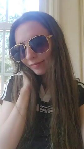 A young woman with long brown hair and sunglasses smiles at the camera. She is wearing a black t-shirt with white writing and trim. She touches her hair and sunglasses. The background appears to be indoors, near a window with trees visible outside.
