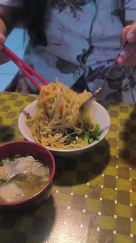 A person in a floral-patterned blouse sits at a table with a yellow and green checkered pattern. In front of them, there is a bowl of noodles with green vegetables and a bowl of soup with dumplings. The person uses red chopsticks in their left hand and a spoon in their right to lift the noodles out of the bowl.