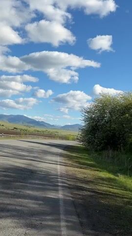 The video shows a road with a vehicle driving on it on a sunny day. The sky is blue with white clouds. On either side of the road there is green grass and trees. In the distance, mountains can be seen.