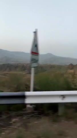 The video shows a view of a hilly, mountainous landscape from the perspective of a moving vehicle. A white and black guardrail is visible in the foreground, blurring due to motion. The landscape consists of rolling hills covered in dry grass and scattered vegetation. In the distance, larger mountains are visible, shrouded in a light haze. A winding road can be seen traversing the terrain. The sky is overcast and pale, suggesting either early morning or late evening light. There are no people or animals visible in the video. The overall impression is one of a natural, somewhat arid environment observed during travel.