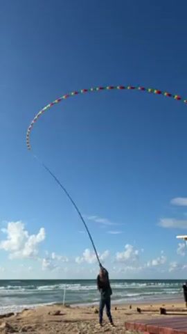 A person is flying a long, segmented kite on a beach. The kite is colorful, and the person is using a fishing pole to control it. The sky is blue with a few clouds. The ocean is visible in the background. The person is wearing dark pants and a jacket.