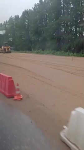 The video shows a construction site with several pieces of heavy machinery. In the foreground, there is a white plastic barrier. Further back, there are two yellow bulldozers and a yellow front-end loader. One of the bulldozers is a large crawler bulldozer, and the other appears to be a smaller, wheeled bulldozer. The front-end loader has its bucket raised. There is also a large road roller or compactor, which is also yellow. In the background, there are trees and a cloudy sky. The ground is covered in dirt or sand, and there are tire tracks visible. Some black pipes are stacked on the ground. The video appears to have been filmed from inside a vehicle, as evidenced by the reflection on the glass and the slight motion blur. The weather appears to be overcast or rainy.