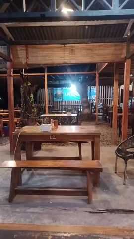 The video shows a casual outdoor or semi-outdoor dining area at night. The space is illuminated by overhead artificial lights, casting a warm glow. The foreground features a wooden table with two matching wooden benches, one pushed underneath it. The table has the number "12" displayed on its side and a small white box on top. To the right, a black metal chair with a woven back is visible.

The dining area has a rustic feel, with a wooden post-and-beam structure supporting a corrugated iron roof. Woven bamboo blinds are rolled up, hanging from the roof structure. The ground beneath the tables is a mix of concrete and gravel or dirt.

In the background, more tables and chairs are visible, along with some trees or large plants. A bright blue sign, partially obscured and difficult to read, appears to be for "J.M. Kopi" or a similar establishment, suggesting it's a coffee shop or eatery. A man in a light-colored shirt walks from the right side of the frame towards the left, passing through the main seating area. The overall atmosphere appears relaxed and quiet, typical of an evening setting in Geulumpangmeujim-Jim, Indonesia.