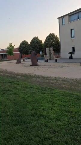 The video pans across a park-like area with a building in the background. The building appears to be a modern structure with light-colored walls and large windows. In front of the building, there are several large stones that seem to be part of a sculpture or landscaping. The ground in front of the building is a mix of sand and gravel, and there is a grassy area in the foreground. Several trees are visible, and the sky is a light blue, suggesting it is daytime. There are no people clearly visible or interacting in the scene.