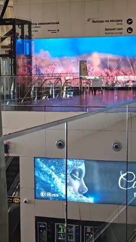 The video captures an interior view of what appears to be an airport or a large public building. On the upper level, a large LED screen displays a scenic image of cherry blossoms against a blue sky with clouds and birds. Below this, a digital advertisement shows a woman's profile with a frosty, icy effect, accompanied by the brand name "Russ." There are escalators visible, along with people walking and sitting. Another digital screen displays text that is partially obscured. Further down, there is a structure resembling a small market stall with merchandise, and people are seen browsing. The architecture features concrete pillars and large glass windows. Signs in Russian indicate "Airlines" and "Exits to Gates/Business Lounge."