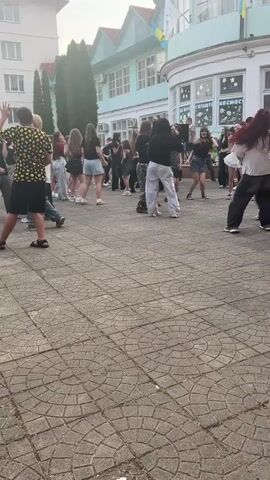 A group of young people are dancing outside a building on a paved area. The building has multiple stories and appears to be a school or institutional building, with windows and flags visible. Some of the young people are wearing casual clothing such as t-shirts, shorts, and pants. They are arranged in a loose circle, with some individuals dancing more energetically than others. The lighting suggests it is daytime, possibly late afternoon, with some shadows cast on the pavement. The paved area is made of interlocking bricks arranged in a circular pattern. In the background, there are trees and other parts of the building. The overall atmosphere appears to be one of casual social gathering and celebration.
