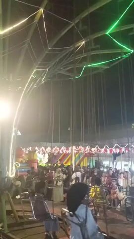 The video shows a crowded fairground at night. Lights illuminate the scene, with a large star-shaped structure adorned with green and white lights overhead. Below, people are gathered, some sitting in what appear to be ride seats. In the background, a Ferris wheel-like structure is visible, as well as various stalls and tents. The atmosphere is festive, with many people present.
