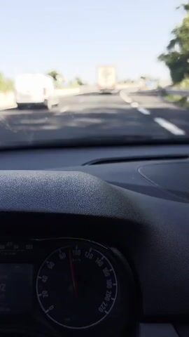 The video shows a POV shot from inside a car, looking out the windshield at a highway. The road ahead is paved and has white lane markings. Several vehicles are visible on the highway, including a white van and two trucks. The sky is clear and blue. The car appears to be in motion.