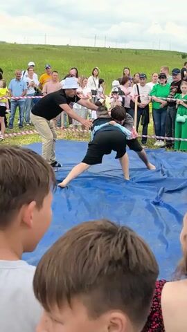 In an outdoor grassy area, a wrestling match is taking place on a large blue tarp. Two young males, wearing black shorts and t-shirts, are grappling. One participant wears a light blue garment over his shoulders and chest. A man in a white bucket hat, black shirt, and khaki pants stands to the side, observing and possibly officiating. He gestures with his left hand, holding a white object. A crowd of spectators, including adults and children, are gathered behind a rope barrier to watch the event. The background consists of a gentle green hillside under a cloudy sky. The camera angle is from behind the spectators, focusing on the wrestling match.