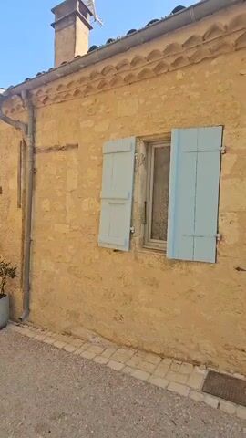 The video shows the exterior of a building with a stone facade. The building has a tiled roof and a gutter system running along the side. There is a window with light blue shutters, which are partially open. To the left of the window, there is another window with glass panes and a view of a plant and some pots. Further to the left, there is a door with glass panels, flanked by potted plants and a yellow mailbox. The ground in front of the building is covered in gravel and bordered by cobblestones. The sky is clear and blue. The camera is panning to the left, showing more of the building's facade and entrance.