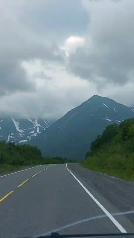 A scenic view from a car as it drives on a paved road through a mountainous landscape. The road is asphalt with a yellow line down the center and a white line on the right edge. On either side of the road, there are green trees and vegetation. In the distance, snow-capped mountains rise, partially obscured by dramatic, grey clouds. The sky is overcast, with patches of lighter grey and hints of blue appearing through breaks in the clouds. The camera is positioned inside the car, capturing the view through the windshield, and a portion of the dashboard is visible in the lower part of the frame. The video appears to be taken during daytime.