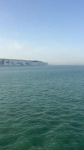 The video captures a serene view of the White Cliffs of Dover from the sea under a clear, pale blue sky. The vast expanse of the turquoise ocean dominates the foreground, with gentle waves rippling across its surface. In the distance, the iconic white chalk cliffs rise majestically, their stark beauty contrasting with the deep blue of the water and sky. The cliffs stretch across the horizon, creating a dramatic natural barrier.

The time of day appears to be morning or late afternoon, indicated by the soft, diffused sunlight that casts a gentle glow on the scene. The weather seems calm and pleasant, with minimal cloud cover. The location is clearly the English Channel, looking towards the coast of Dover, United Kingdom. The overall mood is peaceful and awe-inspiring, emphasizing the grandeur of nature. There are no people or specific objects visible other than the natural landscape and a few distant birds soaring above the water. No text is present in the video.