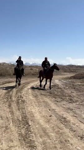 Two men are seen riding horses on a dusty dirt path under a clear blue sky. One man is on a black horse, and the other is on a brown horse. They are moving at a walking pace, casting long shadows on the ground due to the bright sun. In the background, there are mountains, some with snow-capped peaks, and a dry, rugged landscape with sparse vegetation. A red flag is visible in the distance, next to a white sign displaying the Cyrillic text "ШОР-БУЛАК".