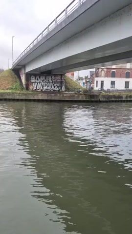 The video shows a body of water, possibly a canal or river, with calm, rippling water. On the left bank, there's a grassy embankment leading up to a concrete retaining wall covered in graffiti. Above the wall, bare trees are visible against a cloudy sky. A bridge, also made of concrete, spans the waterway. The camera appears to be positioned on a boat or other vessel moving along the water. The lighting suggests a gray, overcast day. As the camera moves, more graffiti covered retaining walls come into view along the bank.