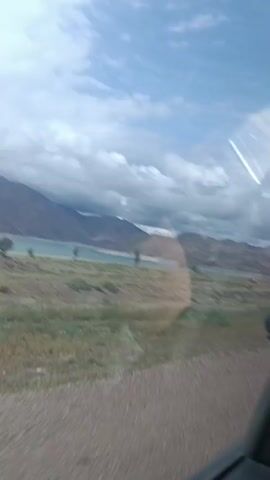 This video appears to be captured from inside a vehicle, looking out through the passenger side window. The view shows a natural landscape with a body of water in the distance, likely a lake, surrounded by hills and mountains. The sky is partly cloudy with patches of blue. In the middle ground, there is a grassy plain with several animals, possibly deer or elk, grazing. The foreground shows a dirt road and the interior of the car, including the car door and a reflection on the window. The camera is in motion, suggesting the vehicle is driving.