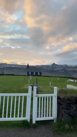 This video captures a serene landscape featuring the iconic Búðir Black Church (Búðakirkja) in Iceland, set against a dramatic mountainous backdrop. In the immediate foreground, a white wooden picket gate is visible, partially open, with a dark, low stone wall beneath it. The scene looks into a verdant green graveyard, where numerous gravestones and white cross markers are scattered across the grass.

The prominent feature is the small, striking black wooden church, distinguished by its white window frames and a small steeple, positioned in the middle ground. Behind the church, a line of low stone walls stretches across the field, separating the church grounds from the broader landscape. Further in the distance, majestic mountains rise, some of their peaks still capped with snow. The sky is filled with clouds, illuminated by warm, soft light in shades of pink, orange, and yellow, mixed with grey and blue, indicating either sunrise or sunset. The overall impression is one of tranquility and natural beauty. No people are visible.