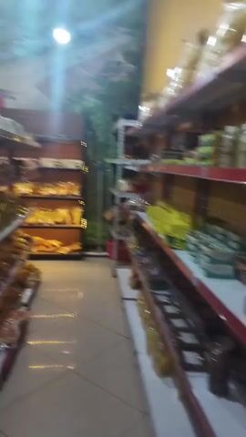 The video pans across a dimly lit, well-stocked grocery store or bakery. Shelves are filled with packaged goods, including what appear to be bread, snacks, and other food items. The lighting is a mix of overhead lamps and accent lighting on the shelves, creating a warm glow. The walls are decorated with a nature-themed mural. The video moves smoothly, suggesting it was filmed by someone walking through the store. The overall impression is of a clean and organized retail space.