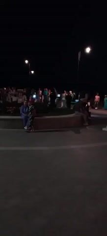The video shows a crowd of people gathered outdoors at dusk or night. Several streetlights illuminate the area, casting a glow on the scene. In the foreground, a curved stone wall or planter box is visible, with a few people sitting on it. A woman in a light blue top and dark pants sits on the left side of the wall, with what appears to be a floral arrangement or wreath nearby. To her right, two people, possibly a man and a woman, are sitting on the wall. The background is filled with a larger group of people, some of whom are standing and talking, while others appear to be observing something or engaged in an activity. The ground is paved with interlocking stones. The sky is dark, indicating it is nighttime.