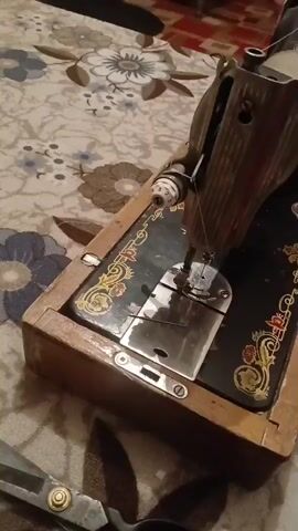 The video shows a close-up view of an antique sewing machine on a patterned fabric surface. Scissors are visible in the lower left corner of the frame. The sewing machine is made of dark metal and wood, with decorative floral patterns on its surface. A person's hand is seen interacting with a piece of white lace fabric. The background is out of focus, but appears to be a room with furniture and other objects. The lighting is dim and appears to be artificial.