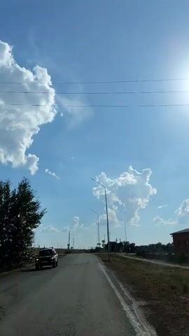 This is a point-of-view video from a vehicle traveling on a paved road. The sky is bright blue with fluffy white clouds and the sun is shining. There are streetlights along the side of the road, and a few signs are visible. On the left side of the road, a dark-colored SUV is visible. The road appears to be in a rural or suburban area, with sparse vegetation and some buildings in the distance. The vehicle is moving at a moderate pace.