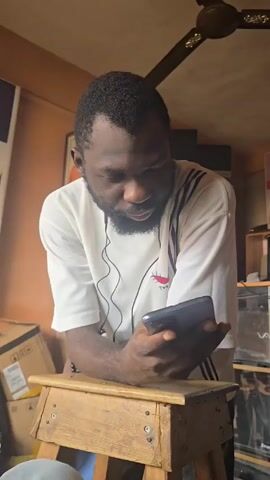 A medium-shot video shows a man with a dark complexion, short black hair, and a beard, wearing a white t-shirt with black stripes on the shoulders. He is leaning on a small, wooden stool, holding a smartphone in his hands, and looking at the screen. He briefly looks up at the camera and speaks. He has earphones connected. The background includes cardboard boxes, a ceiling fan, and electronic equipment on a shelf.