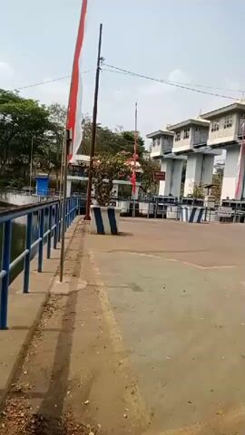 The video shows a body of water with a blue railing along its edge. To the right, there are several white structures with windows, possibly control towers or gatehouses, arranged in a row. These structures are supported by tall white pillars. In front of these structures, there are blue and white striped barriers or bollards. Red and white flags are flying on poles. Trees are visible in the background. The ground in the foreground is a mix of dirt and concrete. The sky is bright and appears to be daytime. There are no people visible in the video.