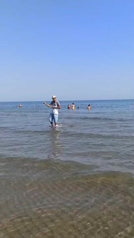 A man wearing a white hat, blue and white t-shirt, and light blue shorts walks through shallow water at a beach. He is holding a fishing rod and casting it out. Other people are in the water and on the beach in the background. The sky is clear and blue. The water is calm with small waves.