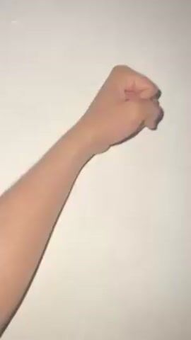 A person's arm and hand are positioned against a plain, off-white wall. The hand starts as a clenched fist, then the pinky finger is extended, and finally the hand is fully opened.