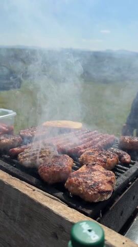 The scene depicts a barbecue grill laden with assorted meats: predominantly sausages and several burger patties. Smoke billows upwards from the grill, obscuring parts of the landscape in the background, which features a green field and a range of hills under a bright blue sky with scattered clouds. A piece of bread is also seen on the grill. To the left of the grill, a plastic container holding food items is visible, along with a green bottle. A person, whose hands are visible, uses tongs to manage the meats.  The grill itself is set on a wooden structure. A car can be seen in the background, further emphasizing the outdoor setting.