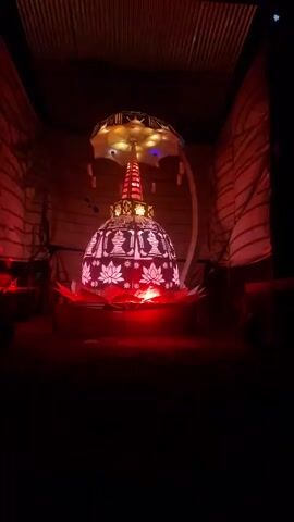 In a dimly lit space, a Buddhist stupa made from lights stands on a red lotus platform. The structure has a dome shape adorned with white lotus designs and figures. A golden spire rises from the top, and an umbrella-like structure is overhead. A curved arch with multicolored lights decorates the scene. The backdrop consists of light-colored fabric draped on walls, with potted plants on either side of the stupa. The space is enclosed by a corrugated roof. Music is playing in the background.