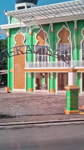 The video pans across the facade of a green mosque with white and orange accents. The mosque has a black dome and intricate geometric patterns on its walls. In the foreground, there is a road with a yellow and black striped barrier. Trees and foliage are visible behind the mosque. The sky is blue.