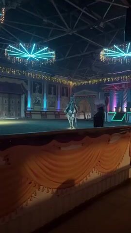 A white horse with a rider dressed in a flowing gown is performing in an indoor arena. The horse is adorned with decorative lights on its harness. The arena has a sandy floor and is decorated with a backdrop resembling ancient Roman architecture with statues and murals. Overhead, circular light fixtures with blue and green lights are suspended, along with strings of small yellow lights. A red carpet leads up to a raised platform on the right side of the arena. In the background, several figures are visible, including what appears to be a person in dark attire standing near the platform. The foreground shows a draped orange fabric structure, possibly a stage curtain or barrier. The lighting is dim, creating a dramatic atmosphere for the performance.